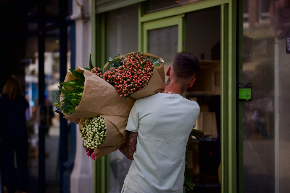 A person with short dark hair, dressed in a white t-shirt, is seen from behind holding a large bouquet of fresh flowers wrapped in brown kraft paper. The bouquet contains pink and white hypericum berr
