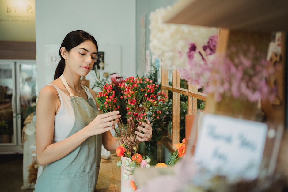 A young woman with dark hair tied back, wearing a white sleeveless top and light grey apron, stands in a flower shop holding a small bouquet of pinkish-red hypericum berries with green stems and leave