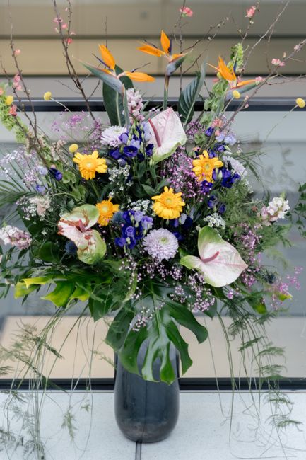 A tall, vibrant floral arrangement placed in a sleek, dark ceramic vase featuring a variety of fresh flowers. The bouquet includes yellow daisies, purple chrysanthemums, white calla lilies with pink s