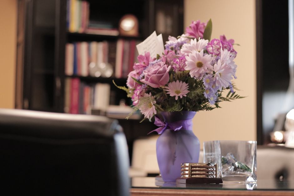 A floral arrangement featuring pink roses, light pink daisies, and purple asters, arranged in a lavender vase with a satin ribbon tied around its neck. The bouquet is placed on a wooden table in an in