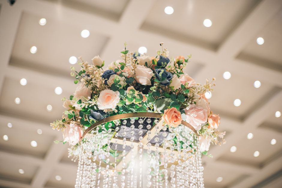 A floral installation featuring a bouquet of soft pink roses, white roses, and blue delphiniums, arranged with lush green foliage and small white accent flowers. The flowers are cascading from a circu