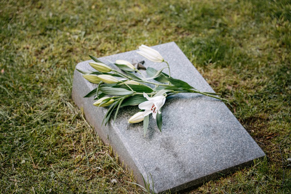 A bouquet of white lilies with closed buds and green leaves rests gently on a flat, grey granite gravestone in a grassy area. The lilies are fresh, with smooth petals and sturdy stems, some flowers pa