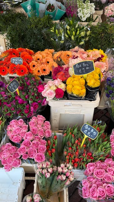 A colorful display of fresh flowers at a market stall, including bright orange gerbera daisies, soft pink roses, yellow tulips, and white lilies, arranged in various containers such as buckets and bas