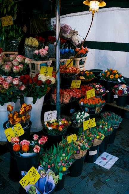 A vibrant flower market stall featuring a variety of floral arrangements and bouquets, including pink roses, multicoloured tulips, and cheerful sunflowers displayed in black buckets and white vases. T