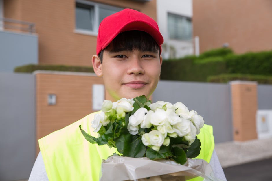 A young male flower delivery courier wearing a red cap and bright yellow high-visibility vest holding a bouquet of fresh white flowers wrapped in white paper. The bouquet features densely packed bloom