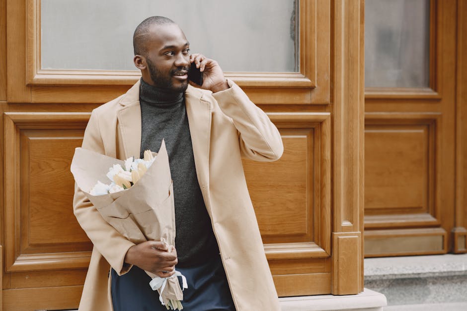 A man with dark skin, a short beard, and a bald head is standing outdoors in front of a wooden-paneled building, holding a bouquet of fresh white and cream-colored flowers wrapped in light brown paper