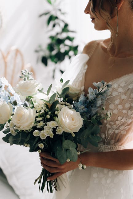 A bride holding a floral bouquet arrangement featuring large white roses, smaller white daisies, and light blue flowers, complemented by green foliage and eucalyptus. The bouquet is wrapped in subtle,
