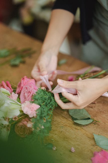 Close-up of a florist's hands wrapping a floral bouquet with soft pink flower arrangements, including roses and ranunculus, accented with green foliage, in pink and cream wrapping paper. The florist i