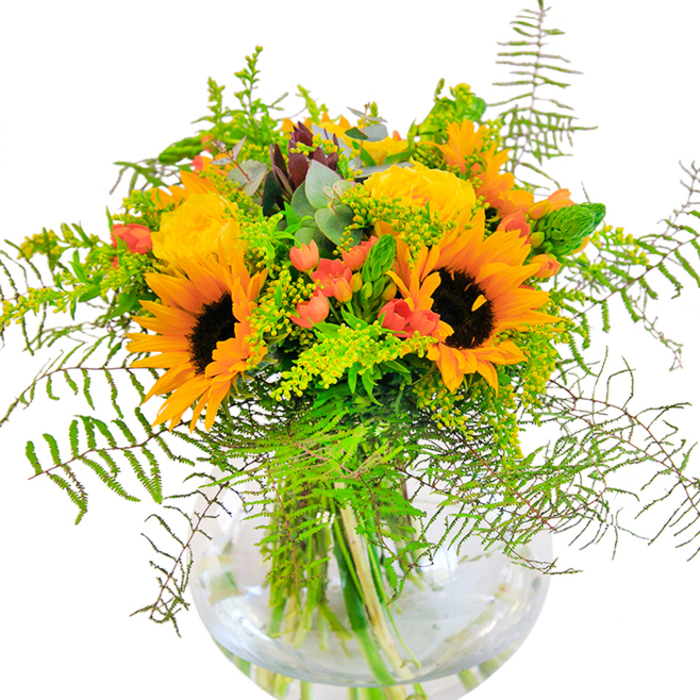 The essence of sunshine is beautifully captured in this eye-catching arrangement from Kingston's local florists. Anchoring the bouquet are brilliant sunflowers, their cheerful yellow and orange petals and textured dark centres instantly recalling the radiant energy of a Kingston summer day. Nestled between, lush yellow garden roses-each petal tightly spiralled-offer a sense of craftsmanship and tradition, their fleecy softness reminiscent of the delicate blooms found in neighbourhood gardens on Lower Ham Road. Drops of red Hypericum berries peek out with unexpected vibrancy, infusing zest and playfulness throughout the ensemble. Lush layers of greenery abound, ranging from trailing feathery ferns to airy spikes of goldenrod, weaving texture and depth that seems almost to rustle in the light, as if caught in a gentle breeze crossing Fairfield Recreation Ground. Subtle notes of eucalyptus, in soft blue-green tones, punctuate the bouquet and add a crisp, invigorating aroma. The entire creation is placed in a round, clear glass vase with a fishbowl silhouette, allowing for the vivid interplay of stems and water to be admired in full view. Bathed in gentle natural light, the arrangement feels both abundant and harmonious-a delightful way to mark an anniversary, birthday, or give cheer to a friend in Norbiton. Every element of this arrangement is a testament to Kingston's distinct floral artistry and local pride.